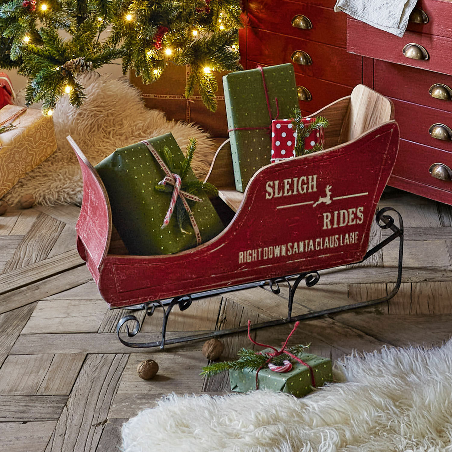Deko-Schlitten Maélys Roter Holzschlitten mit Metallkufen und grünen Geschenken auf Holzboden neben geschmücktem Weihnachtsbaum.