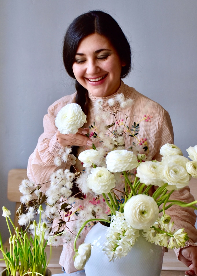 Frau in pastellfarbener Bluse arrangiert weisse Blumen in Vase vor hellem Hintergrund