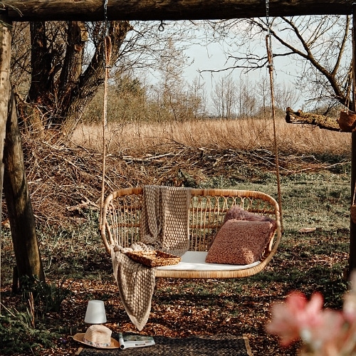 Rattanschaukel in Naturfarbe mit Kissen und Decke vor herbstlichem Gartenhintergrund