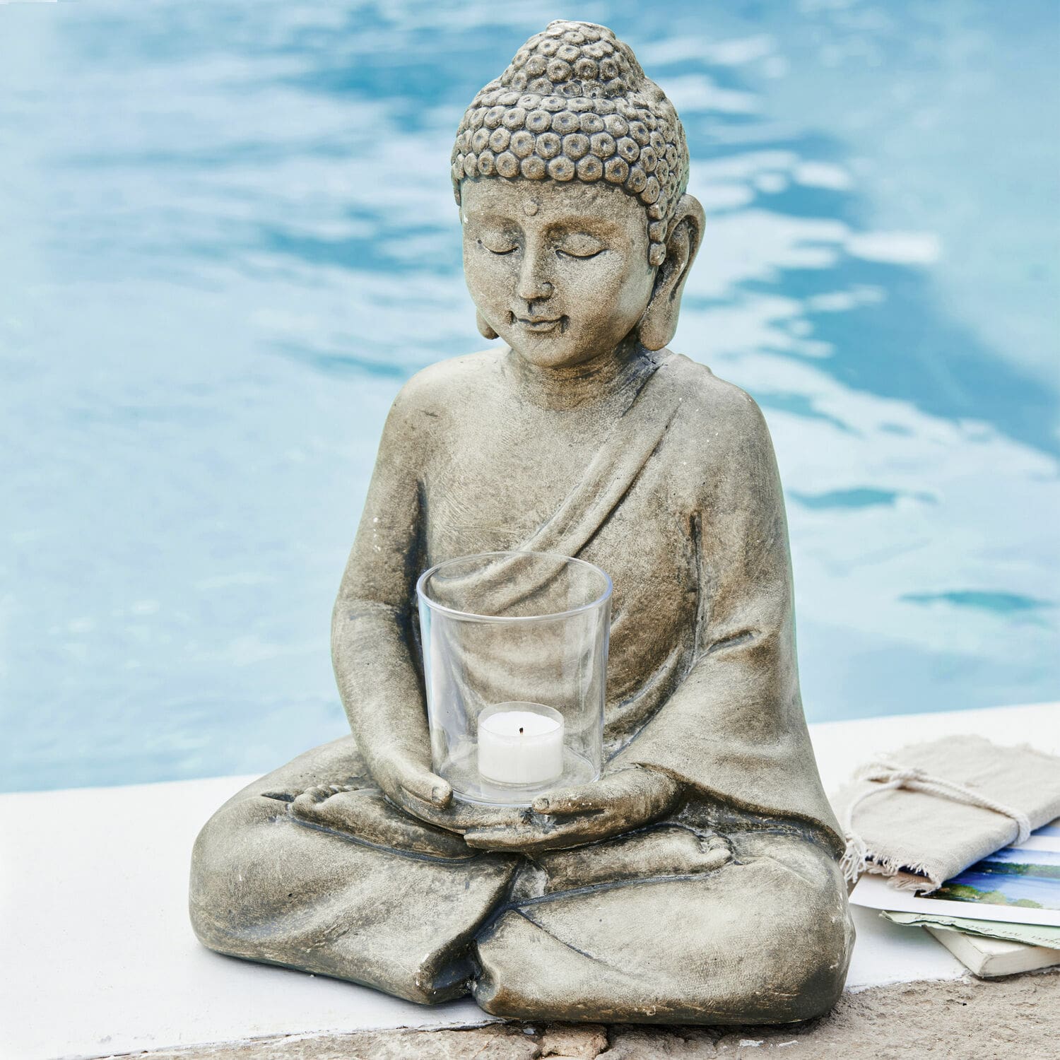 Windlicht Sylvain Graue Buddha-Figur aus Stein mit klarem Teelichthalter am Poolrand für eine entspannte Atmosphäre