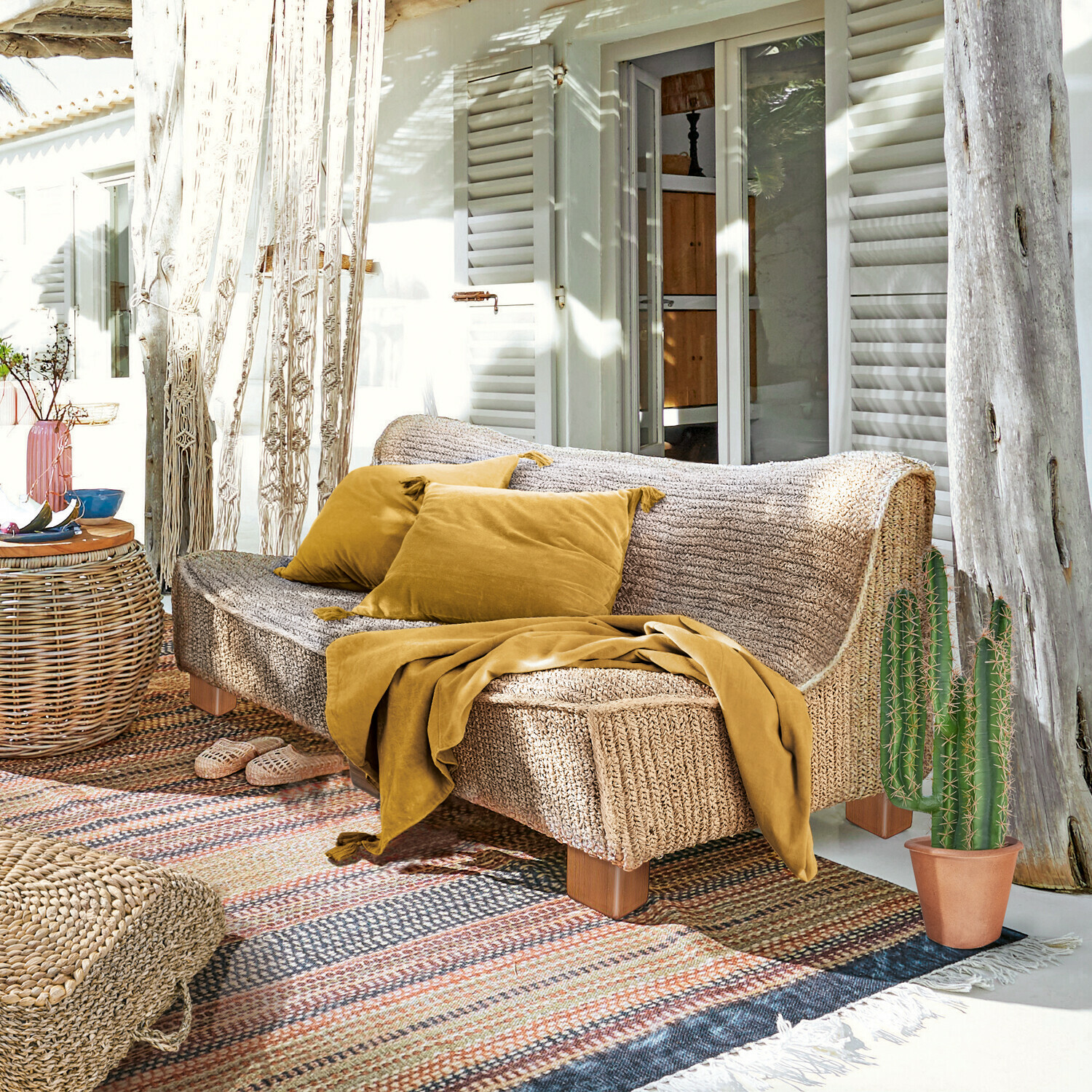 Sofa Bocage Sofa aus geflochtenem Material in Naturfarbe mit senffarbenen Kissen und Decke auf der sonnigen Terrasse