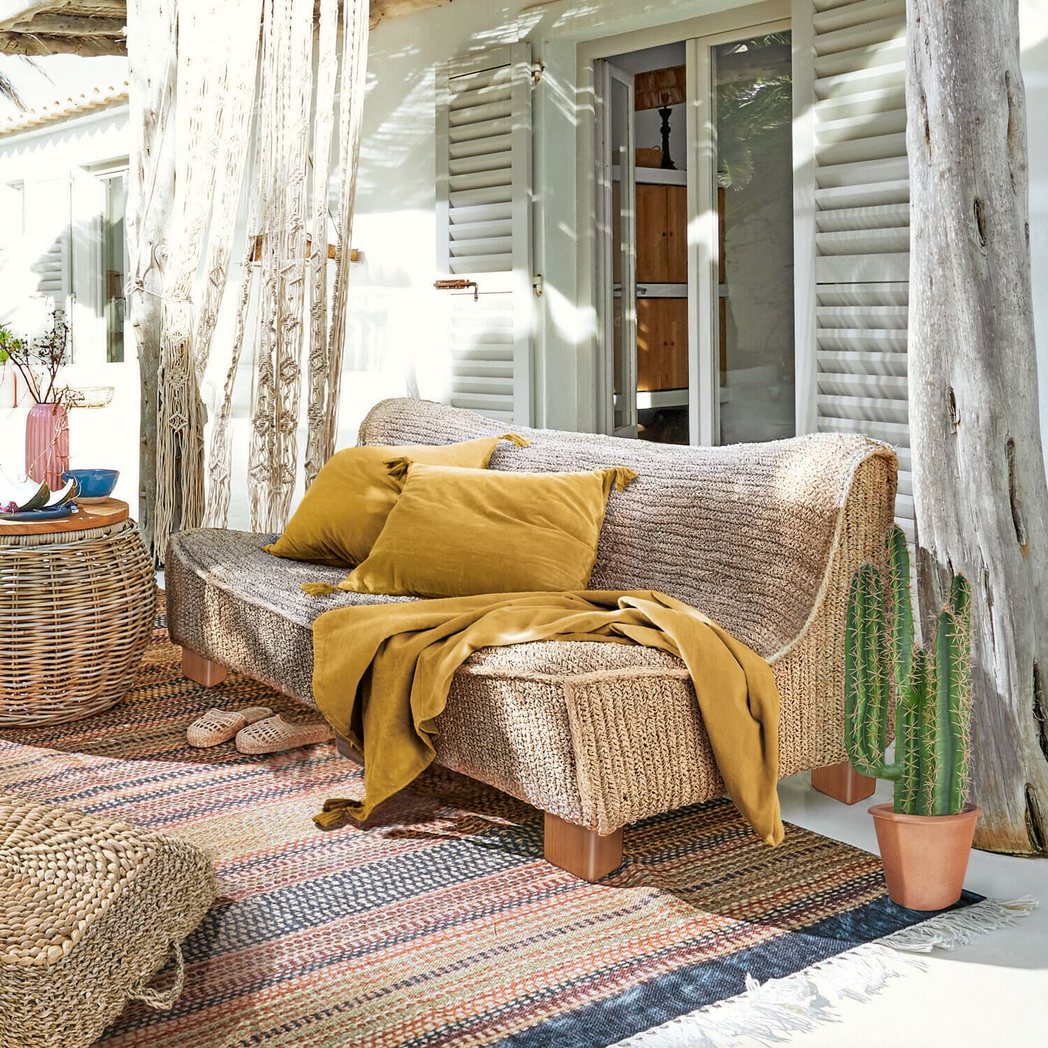 Sofa Bocage Sofa aus geflochtenem Material in Naturfarbe mit senffarbenen Kissen und Decke auf der sonnigen Terrasse