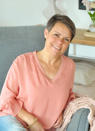Lächelnde Frau in rosa Bluse sitzt auf grauem Sofa in hellem Raum.