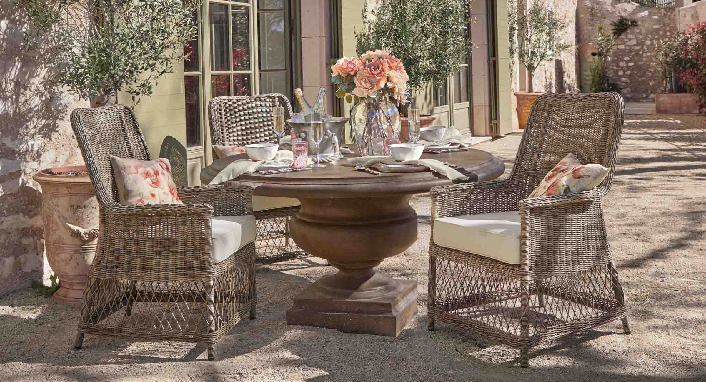 Runder Gartentisch aus braunem Holz mit vier Rattanstühlen und hellen Kissen, mit Blumen dekoriert