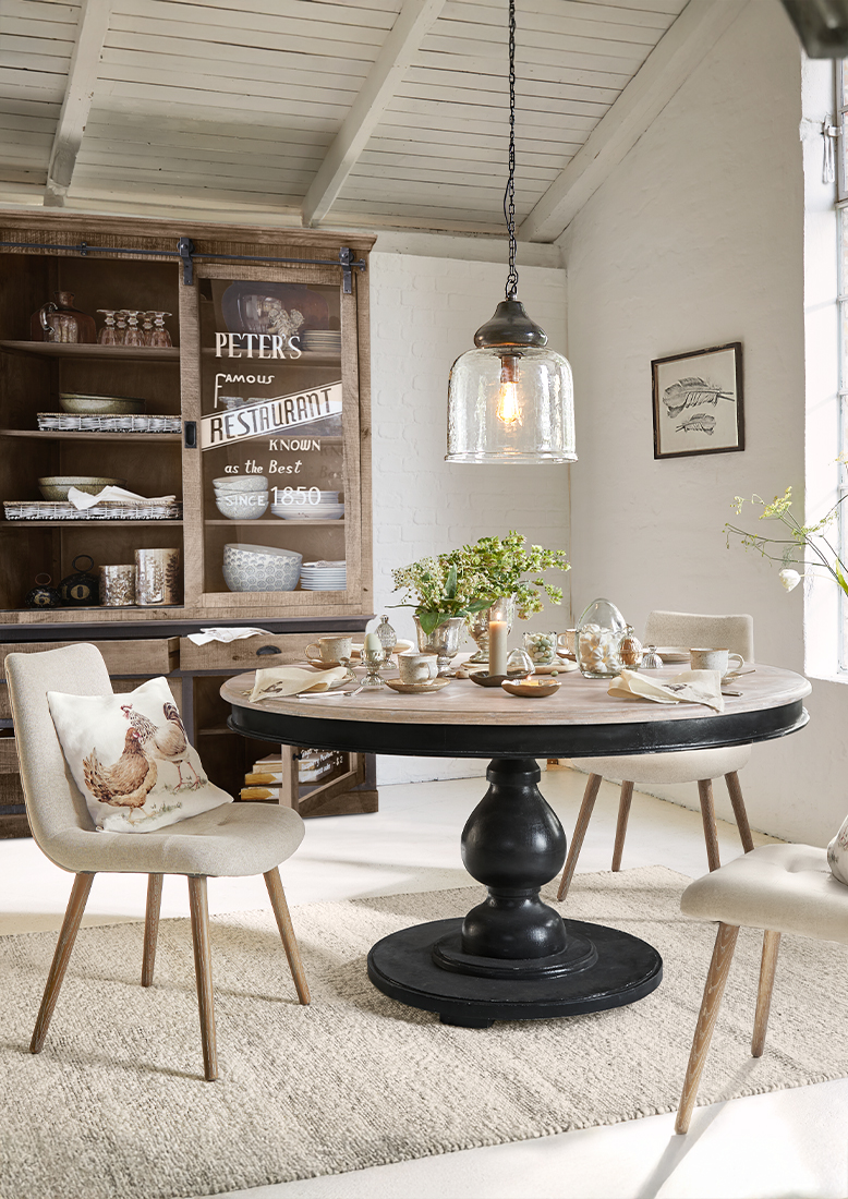 Esszimmer mit rundem Holztisch auf schwarzem Sockel und beigen Stühlen mit Kissen im Hühnerdesign.