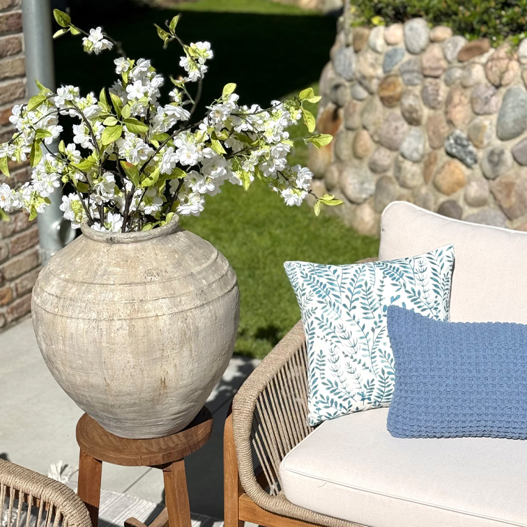 Große beige Keramikvase mit weißen Blumen auf Holzständer neben Sofa mit Kissen im Garten