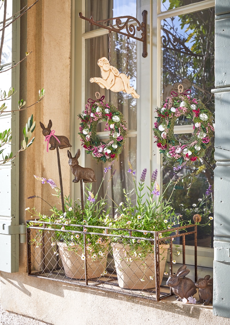 Fensterdekoration mit Metallhaltern, Blumentöpfen und Frühlingskränzen mit Eiern und Hasenfiguren