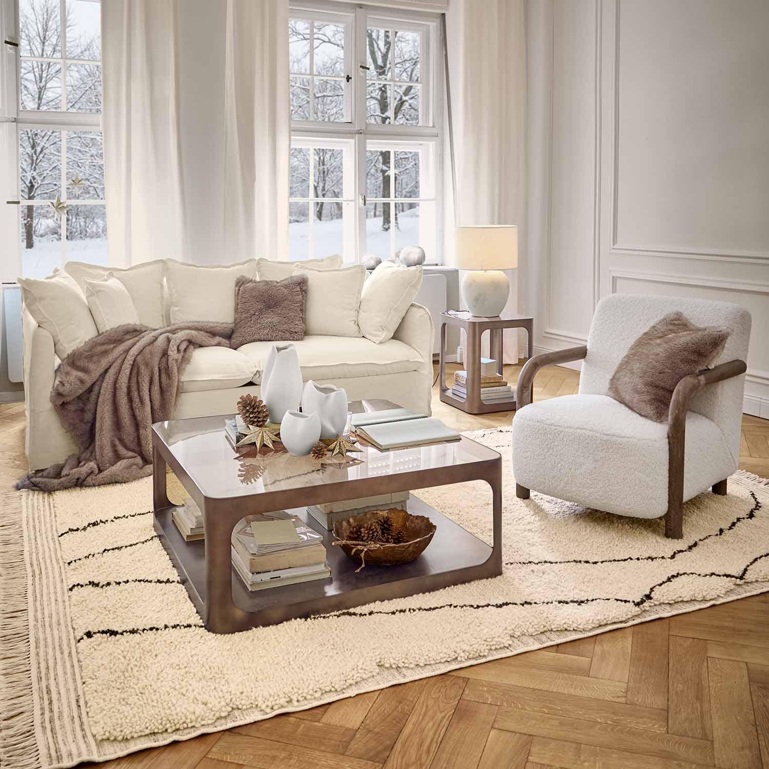 Gemütliches Wohnzimmer mit weissem Sofa, Sessel mit Holzarmlehnen und weichem Teppich im Winterambiente