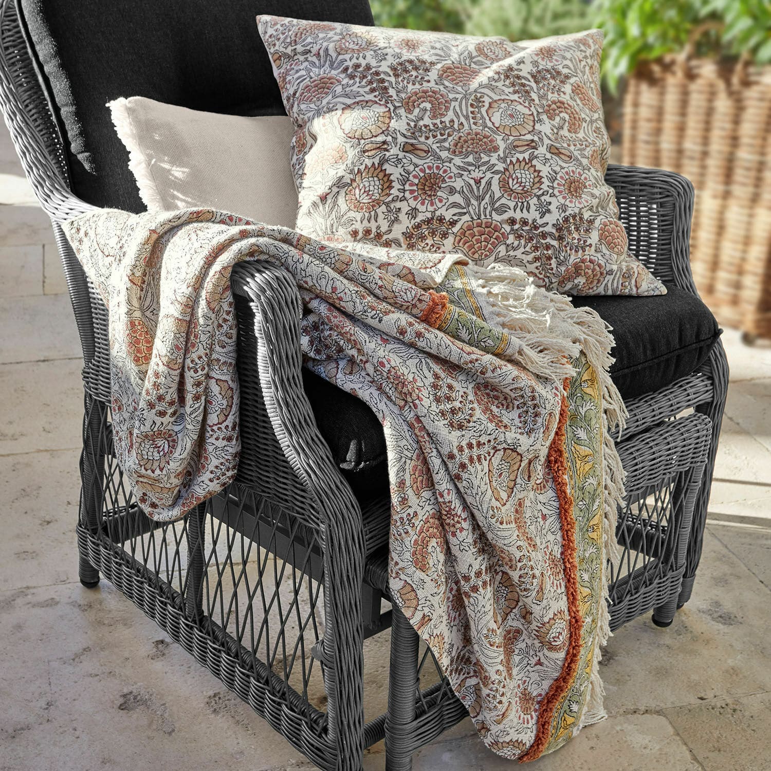 Plaid Romila Baumwolldecke und Kissen mit warmem Blumenmuster auf grauem Rattansessel für eine gemütliche Atmosphäre