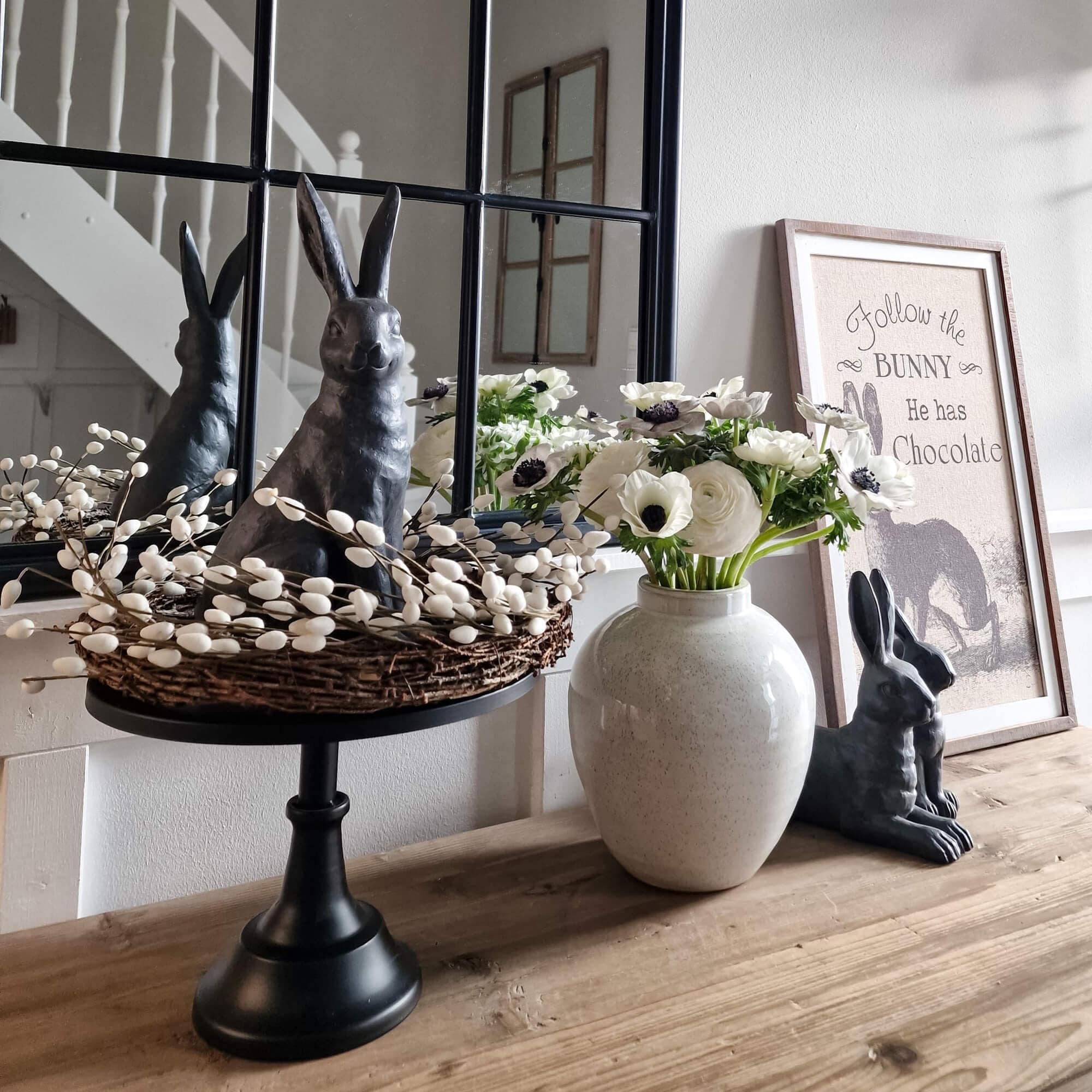 Dekorativer schwarzer Hase mit Weidenkranz und weissen Blumen in Keramikvase auf Holztisch