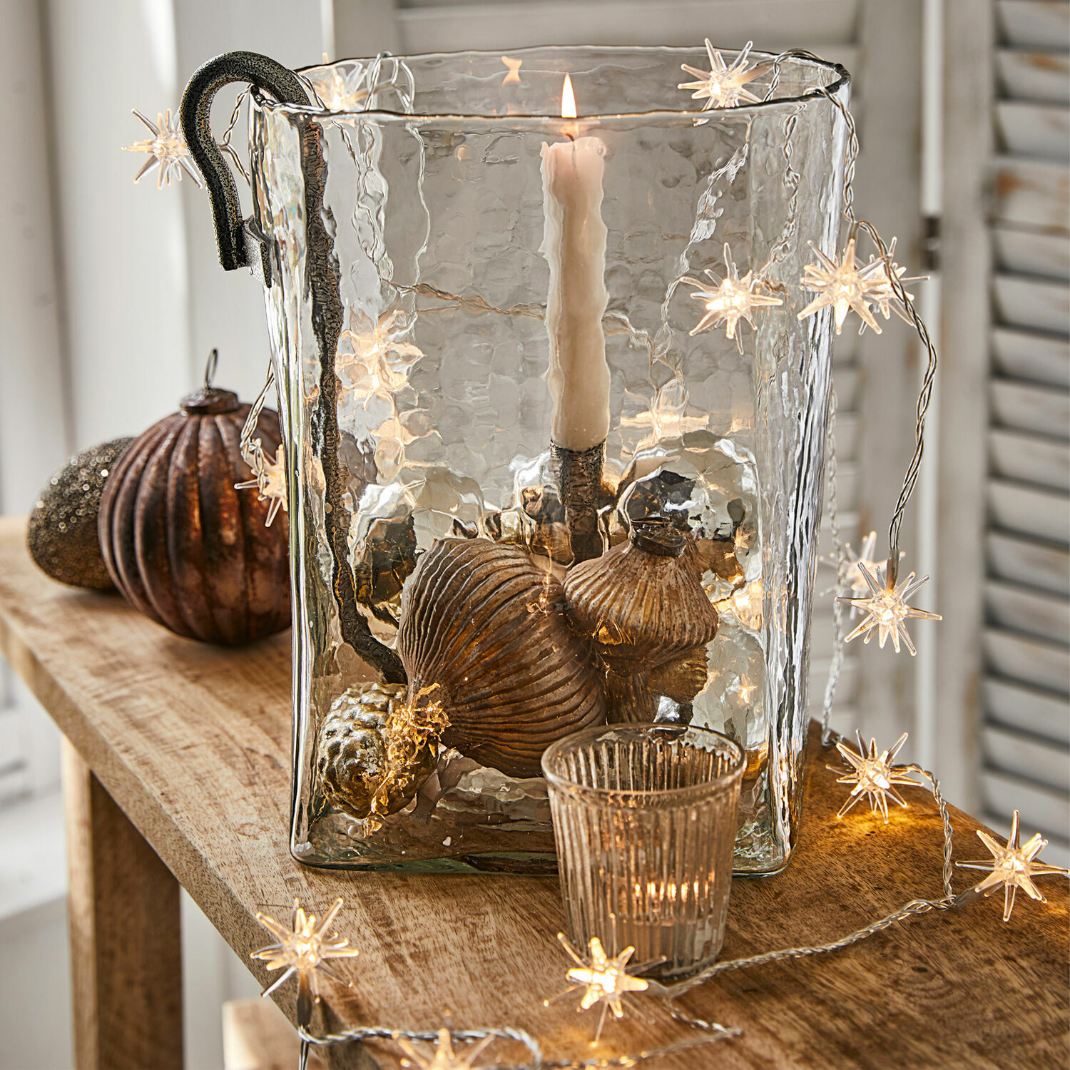 Lichterkette Nelisse Glas-Windlicht mit Metallgriff und kupferfarbenen Dekorationen auf Holztisch mit Sternen-Lichterkette