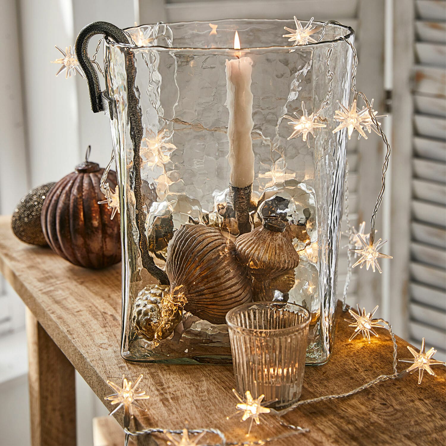 Lichterkette Nelisse Glas-Windlicht mit Metallgriff und kupferfarbenen Dekorationen auf Holztisch mit Sternen-Lichterkette