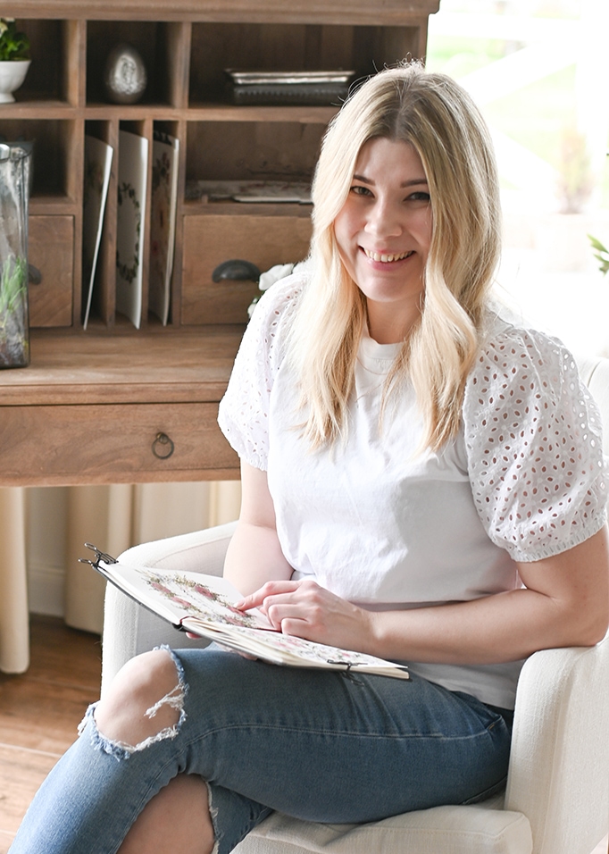 Junge Frau in weisser Bluse mit Lochstickerei und zerrissener Jeans sitzt mit Album in hellem Raum