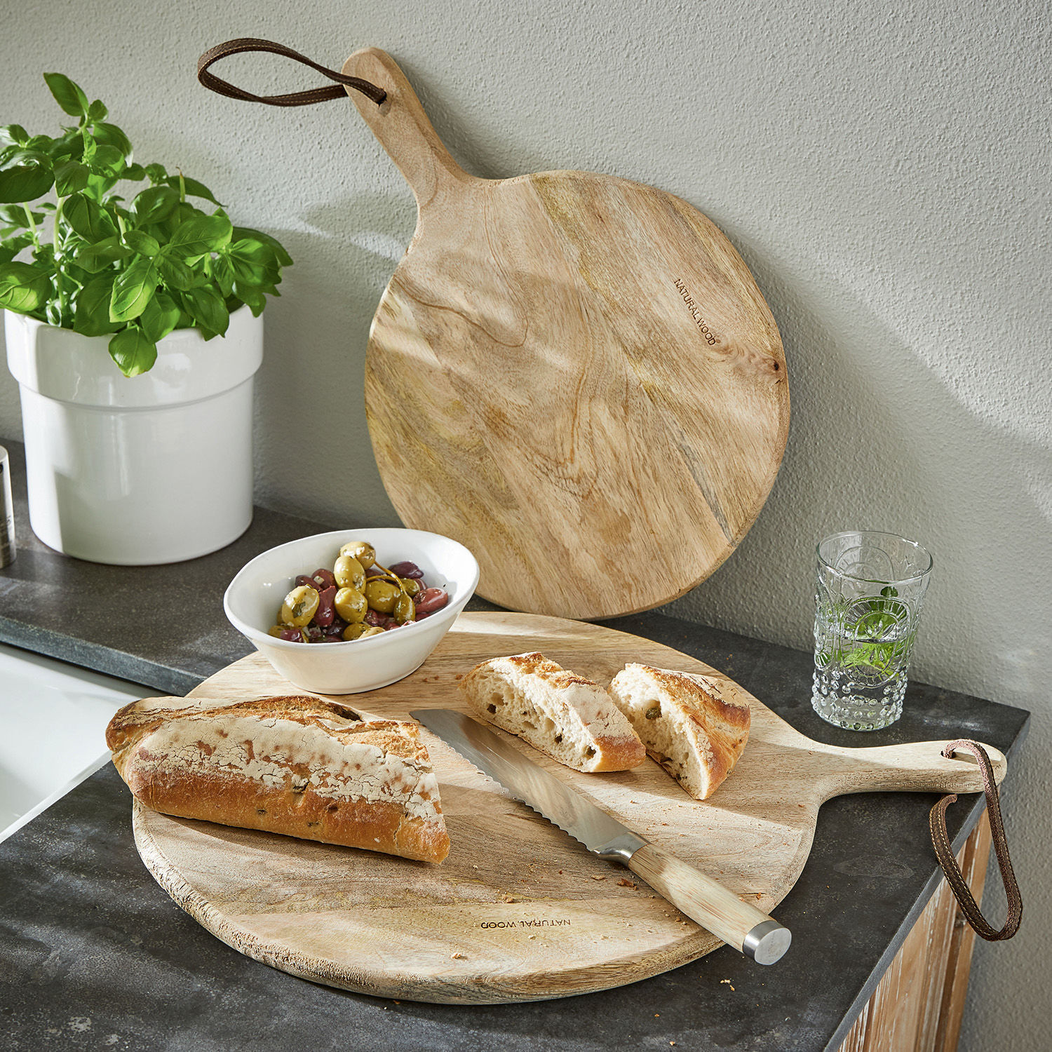 Schneidebrett 2er Set Nitra Rundes Schneidebrett aus Naturholz mit Griff, Brotmesser, Brot und Oliven auf der Küchenarbeitsplatte