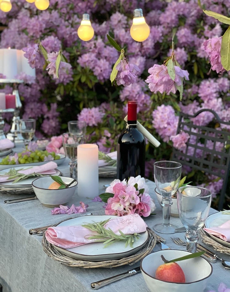 Sommerliche Tischdekoration mit rosa Blumen, Lichterkette und Weinflasche vor lila Blütensträuchern.
