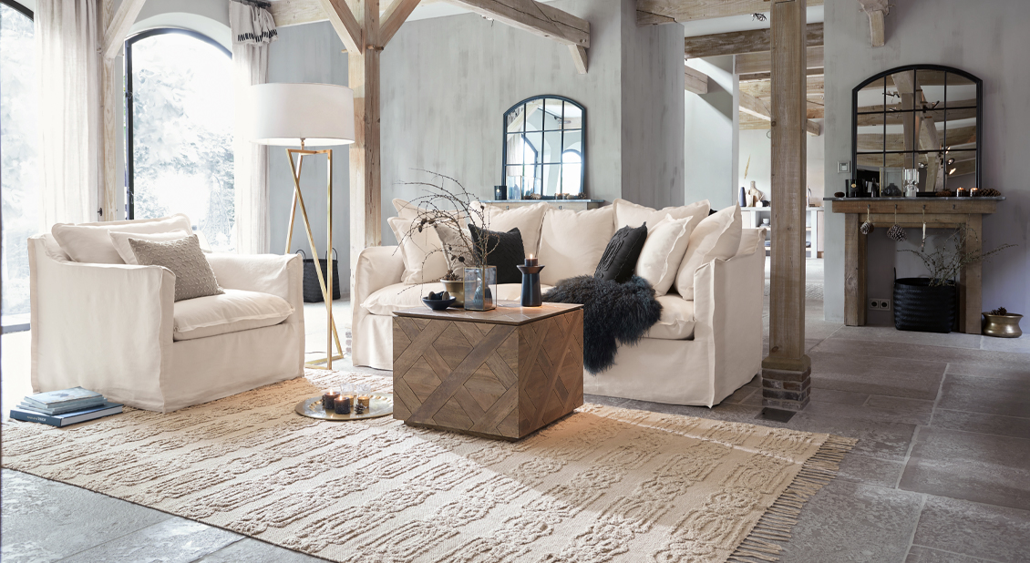Gemütliches Wohnzimmer mit weissem Stoffsofa und Sessel, Holztisch und beigem Teppich im Landhausstil