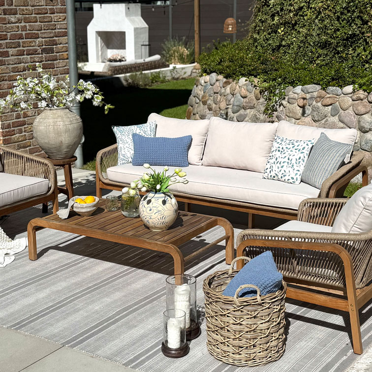 Gartenmöbel-Set mit Holztisch und beigem Sofa mit blauen gemusterten Kissen auf gestreiftem Teppich