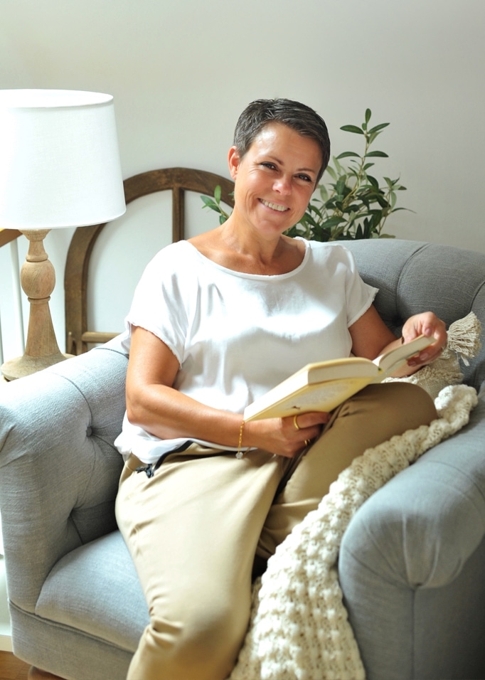 Frau liest Buch auf grauem Polstersessel mit Lampe und Pflanze im Hintergrund.