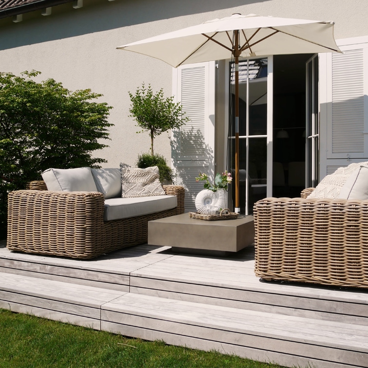 Beiges Rattan-Gartenmöbel-Set mit Sonnenschirm und Kissen auf Holzterrasse im Garten