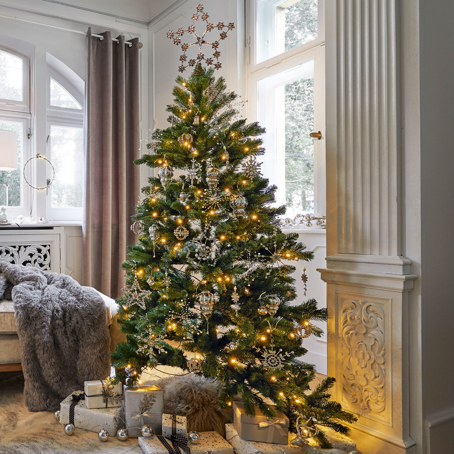 Weihnachtsschmuck-Set Bjouterie Weihnachtsbaum mit grünen Zweigen und silbernen Dekorationen im hellen Wohnzimmer mit Geschenken.