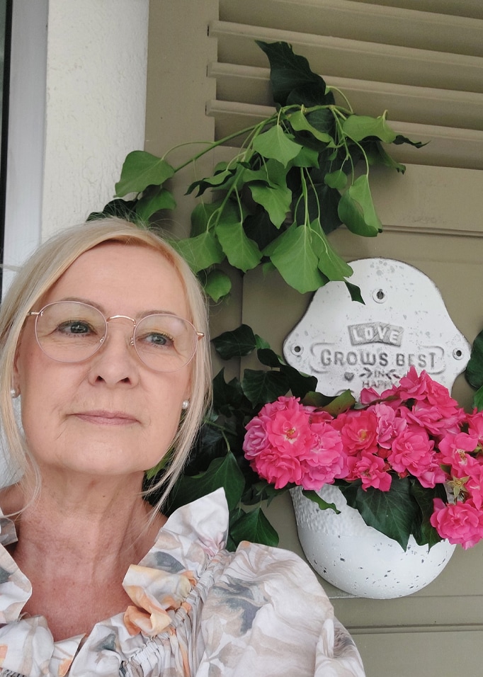 Weisse Wandblumenvase mit pinken Blumen und grünen Blättern vor grauen Jalousien