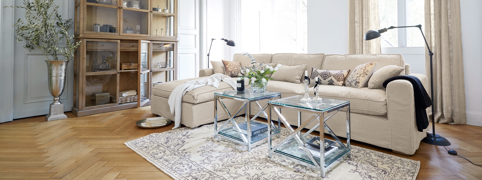 Klassisch elegantes Wohnzimmer mit beigem Sofa, Glastischen und Holzvitrine auf hellem Parkettboden