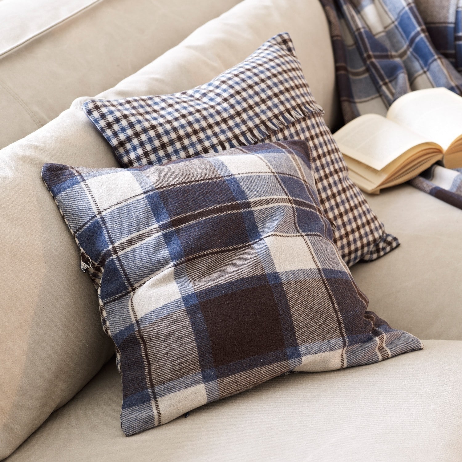 Kissen Clifton jr. Zwei dekorative Kissen mit blau-braunem Karomuster auf beigem Sofa mit offenem Buch.