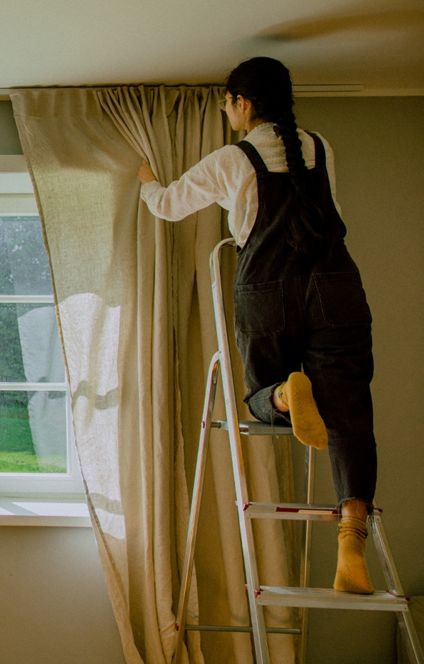 Frau auf Leiter zieht beigen Leinenvorhang vor Fenster in hellem Raum zu.