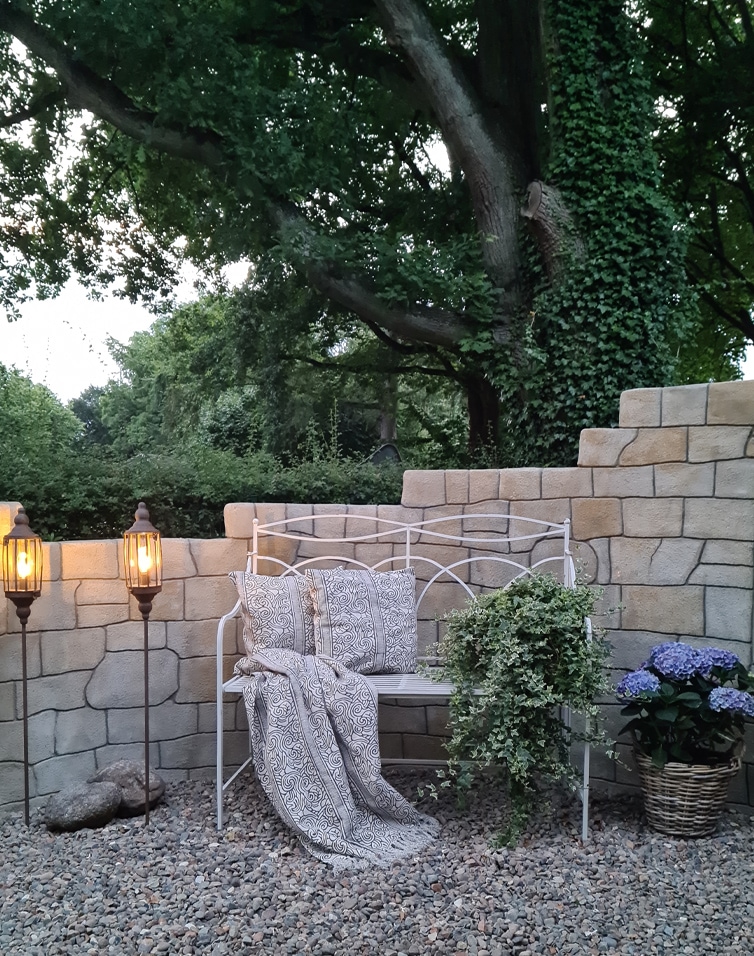 Weisse Metall-Gartenbank mit Kissen und Decke vor Steinmauer, umgeben von Pflanzen und Laternen