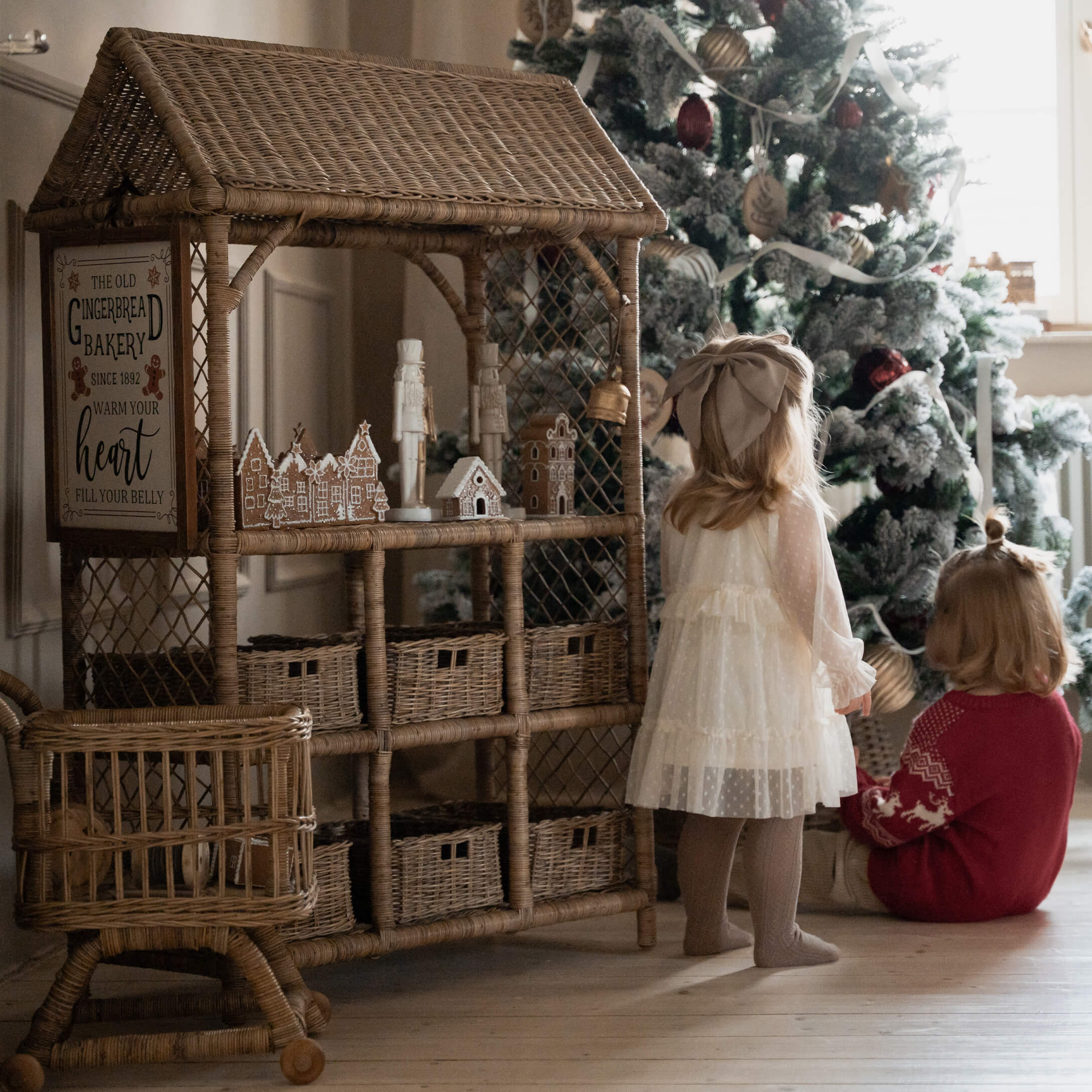 Rattanhaus mit Lebkuchendekorationen neben Kindern vor geschmücktem Weihnachtsbaum