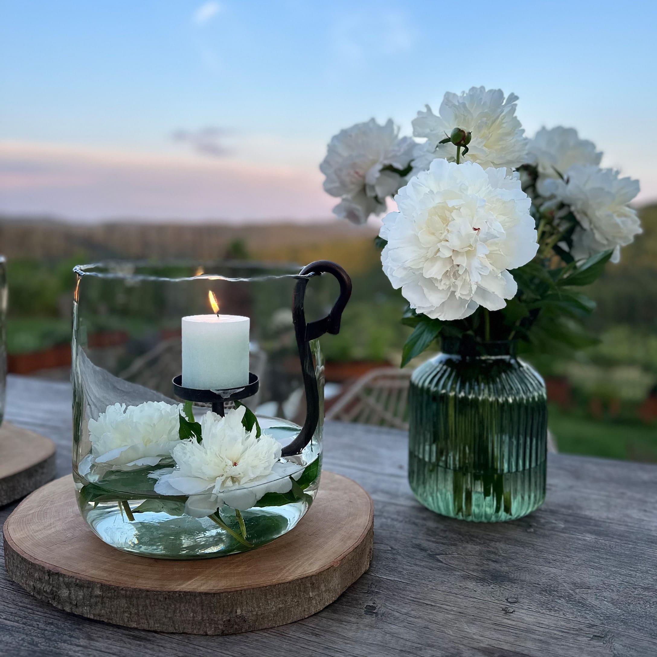 Glaswindlicht mit weisser Kerze und weissen Blumen auf Holzunterlage im Aussenbereich