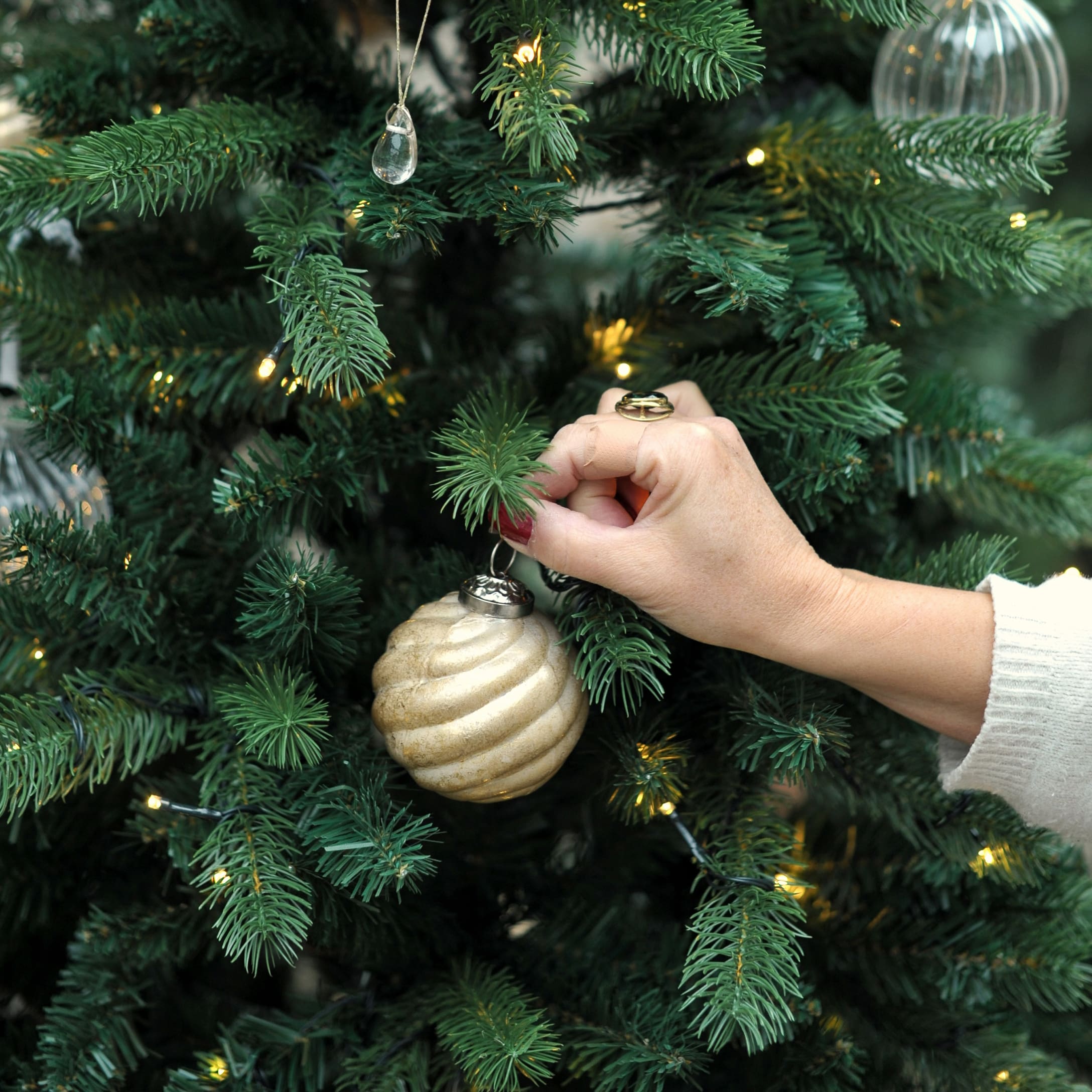 Hand schmückt grünen Weihnachtsbaum mit goldener Spiralkugel und LED-Lichterkette im Wintergarten