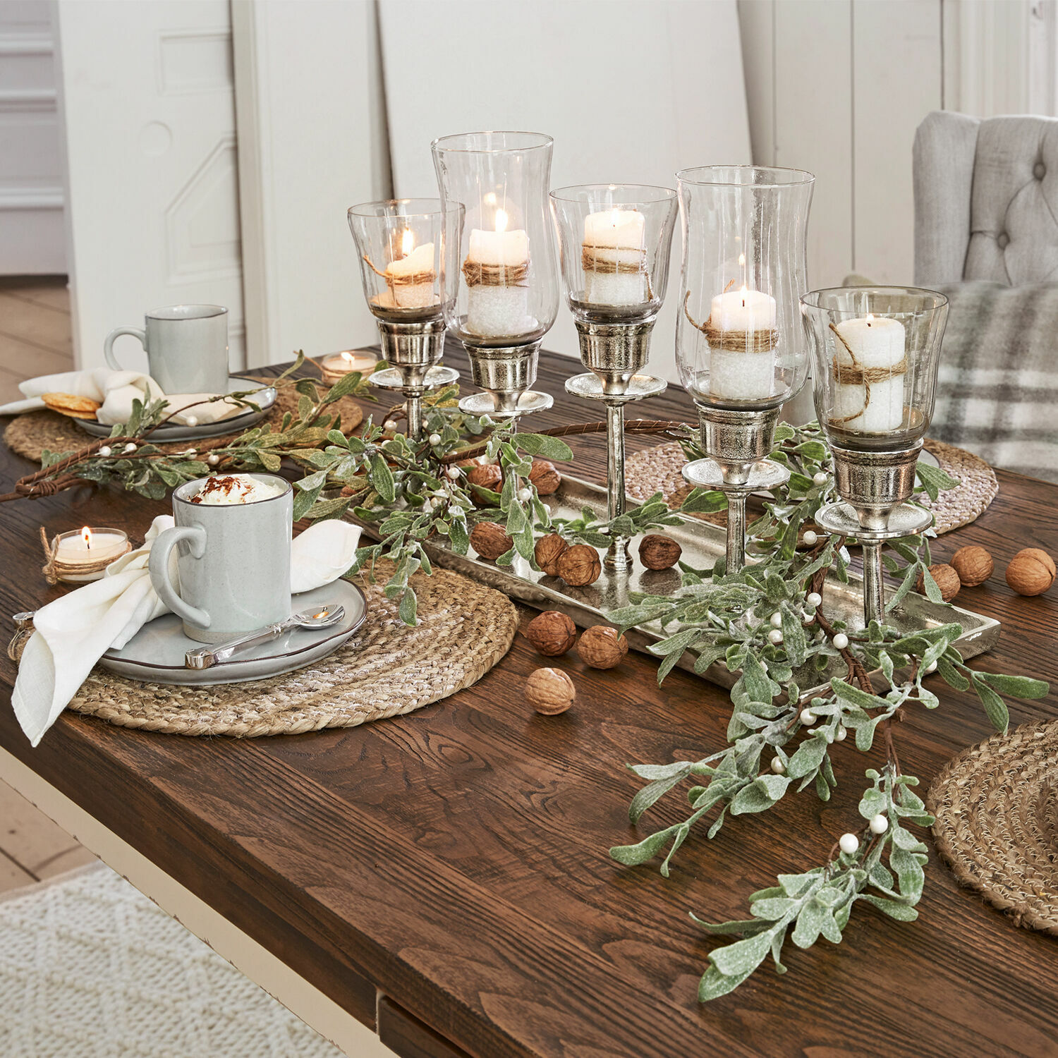 Dekorativer Kerzenhalter aus Glas und Metall auf Holztisch mit grünen Zweigen und Walnüssen.