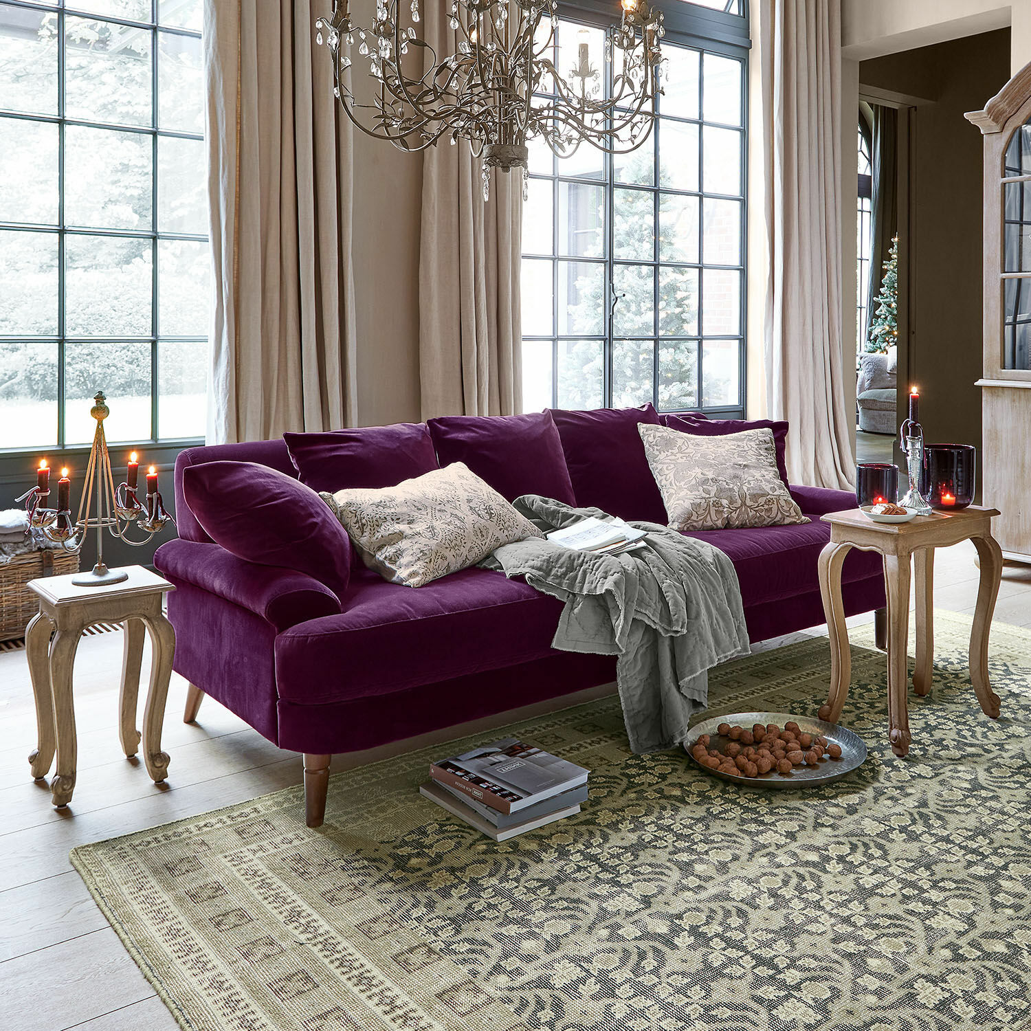 Sofa Rivermere Violettes Samtsofa mit Kissen und grauer Decke in einem hellen Wohnzimmer mit grossen Fenstern und Holztischen