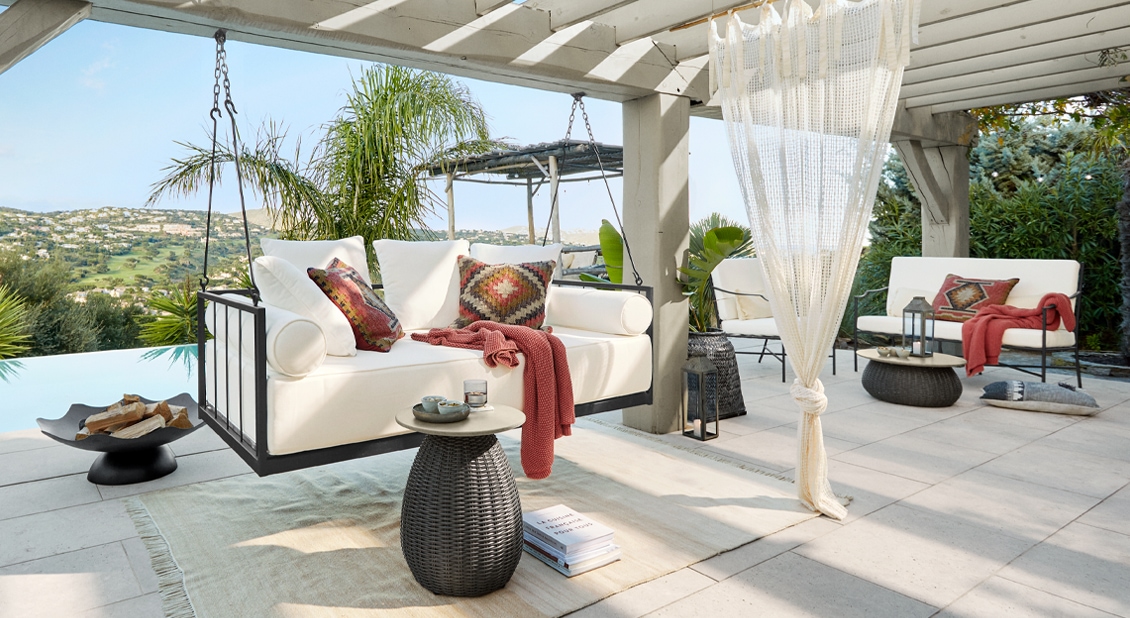 Weisse hängende Sofa mit Kissen und roter Decke unter Pergola auf Terrasse mit grünem Ausblick