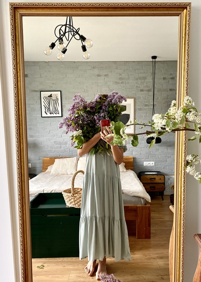 Frau im langen grünen Kleid hält lila Blumenstrauss vor Spiegel im Schlafzimmer mit Holzbett.