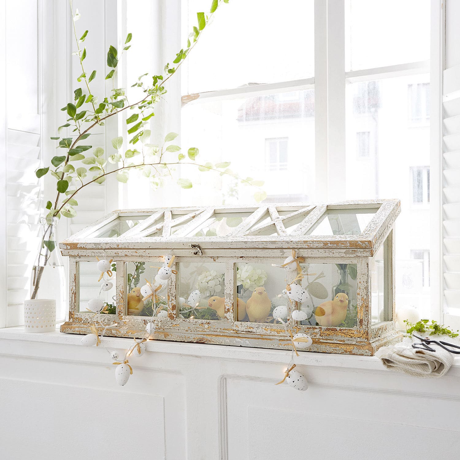 Weisses Mini-Gewächshaus aus Holz im Shabby-Chic mit Küken-Dekoration auf hellem Fensterbrett