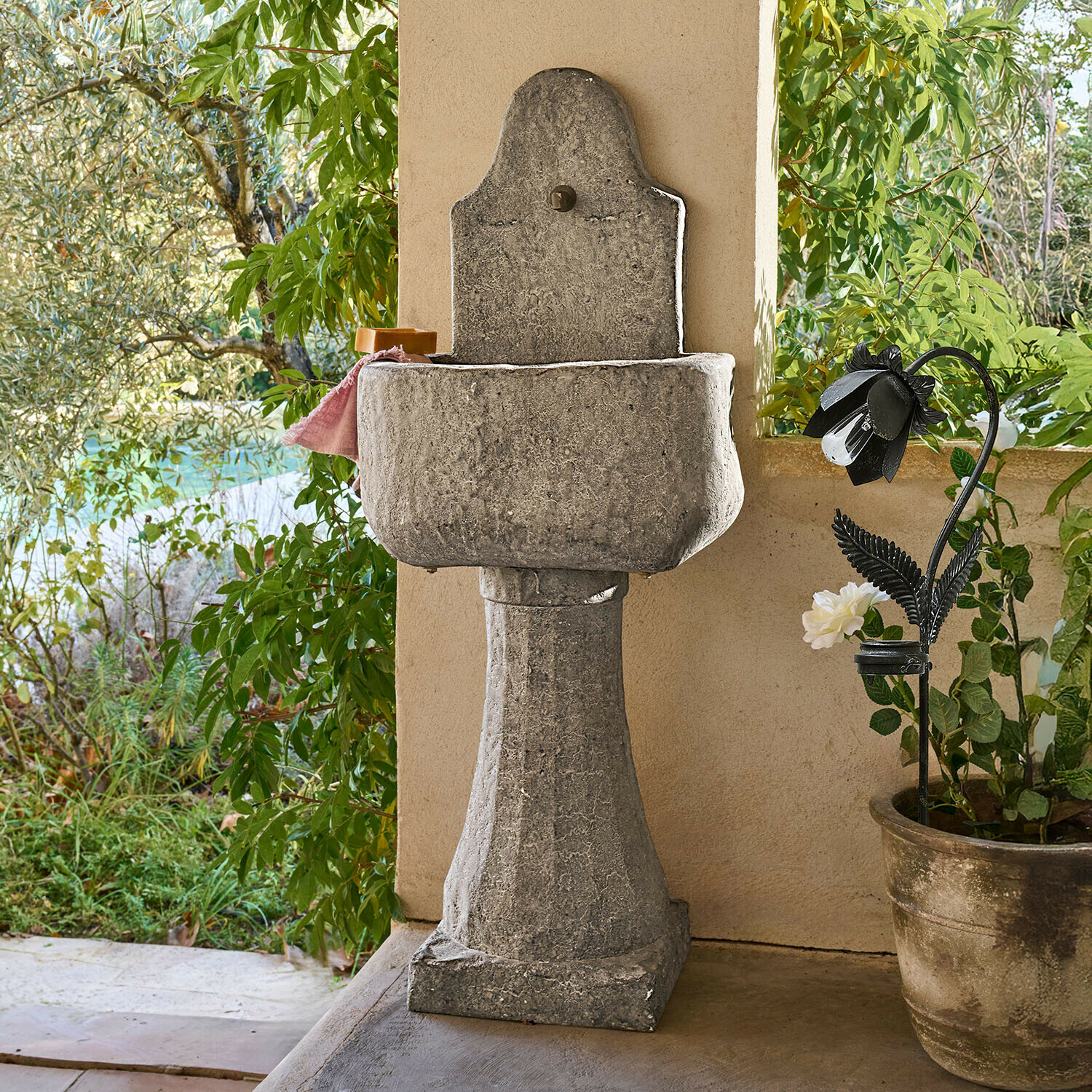 Brunnen Lurevino Dekorativer grauer Steingarten-Waschbecken auf Säule mit natürlicher Oberfläche und Pflanzen im Hintergrund