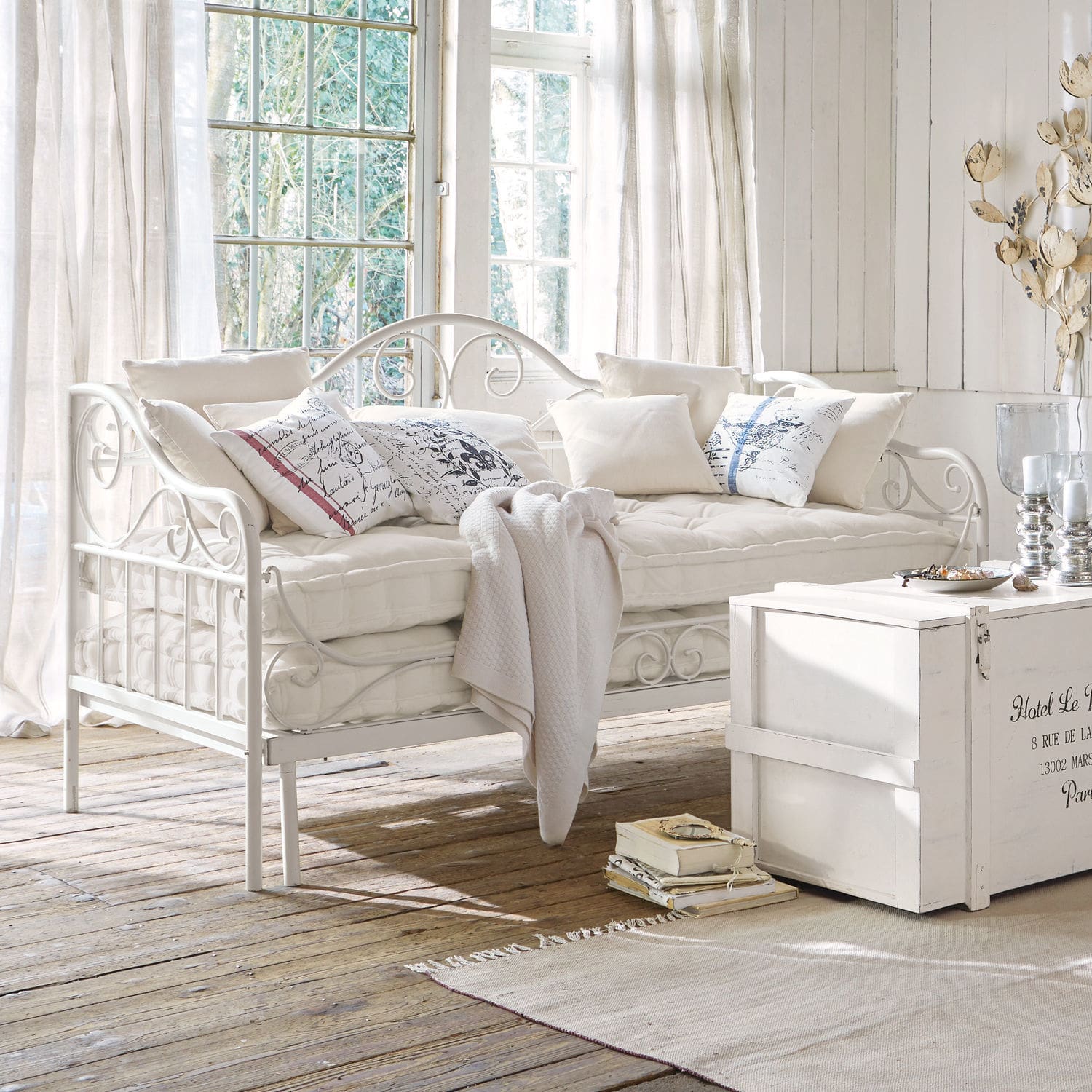 Bettsofa Maditha Weisses Metallsofa mit weichen Kissen und Decke in einem hellen Raum mit natürlichem Holzboden