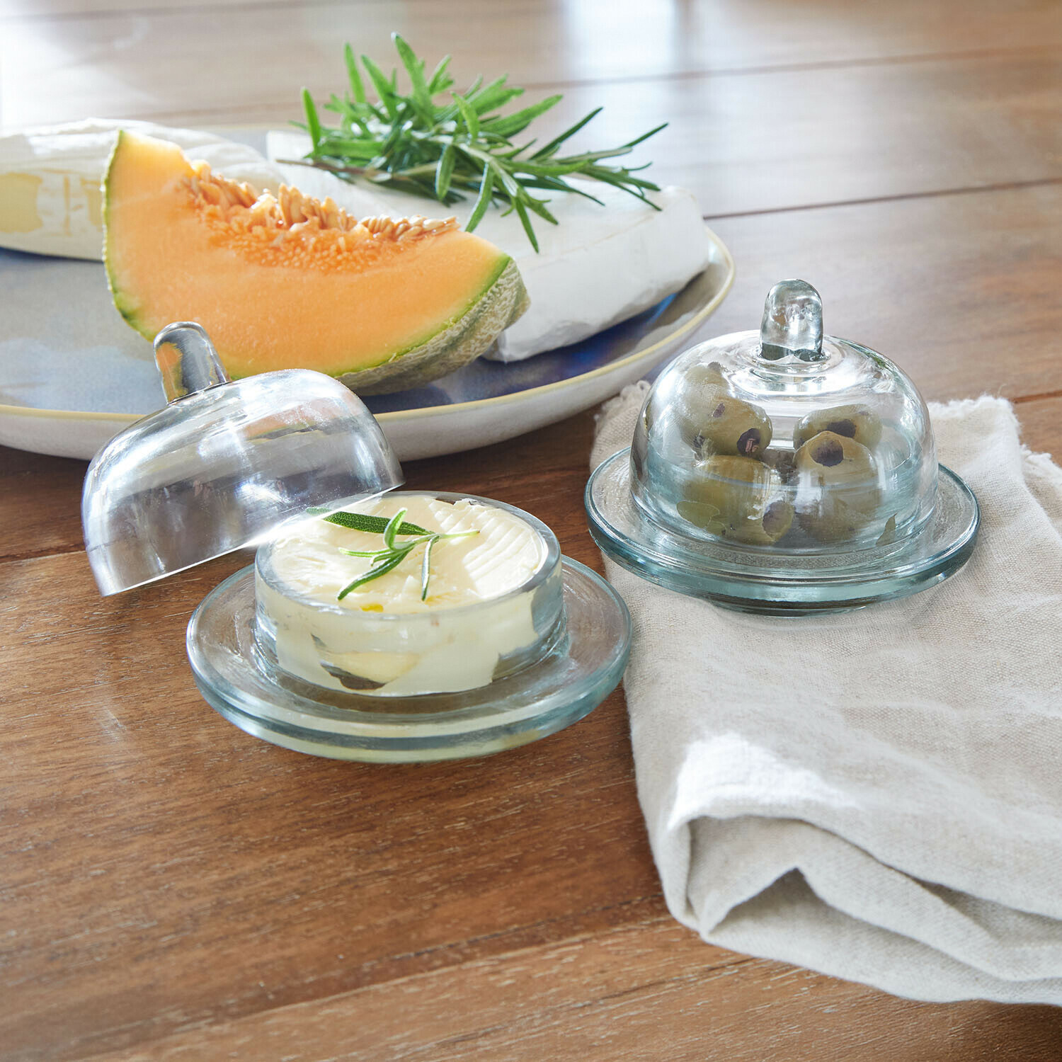 Glasbehälter für Butter und Oliven auf Holztisch mit Melone und Käse im Hintergrund
