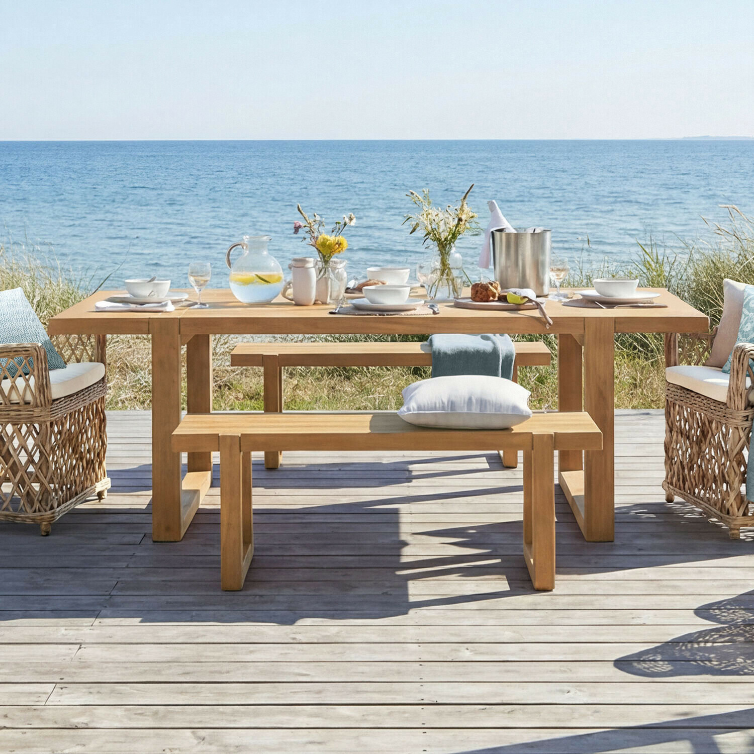 Tisch Halinor Holztisch im Garten mit Bänken und Korbsesseln am Meer, gedeckt mit Geschirr und Blumen