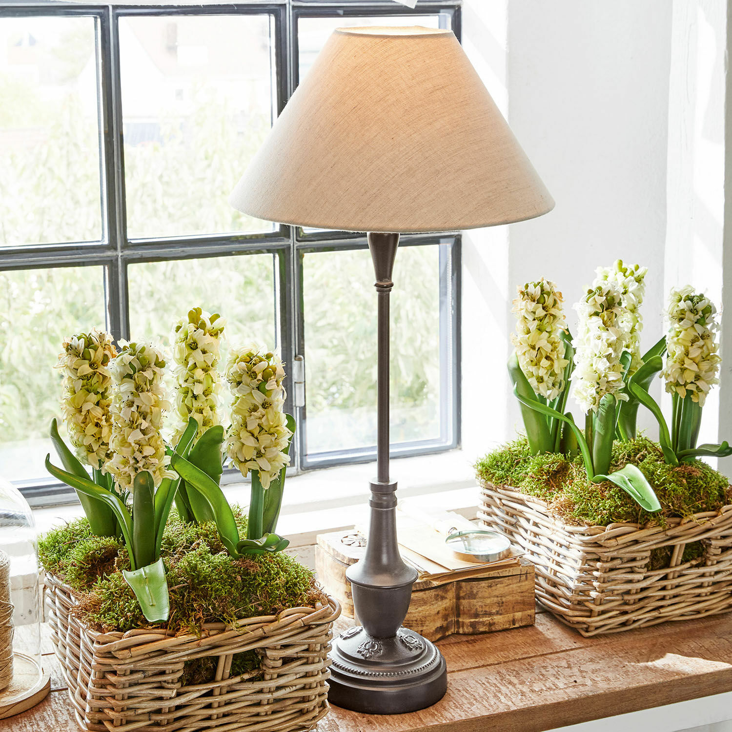 Tischlampe mit Metallfuss und beigem Lampenschirm neben Weidenkörben mit weissen Blumen und Moos