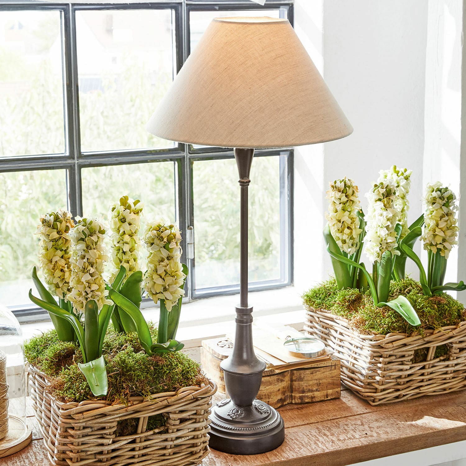 Tischlampe Barnwell Tischlampe mit Metallfuss und beigem Lampenschirm neben Weidenkörben mit weissen Blumen und Moos