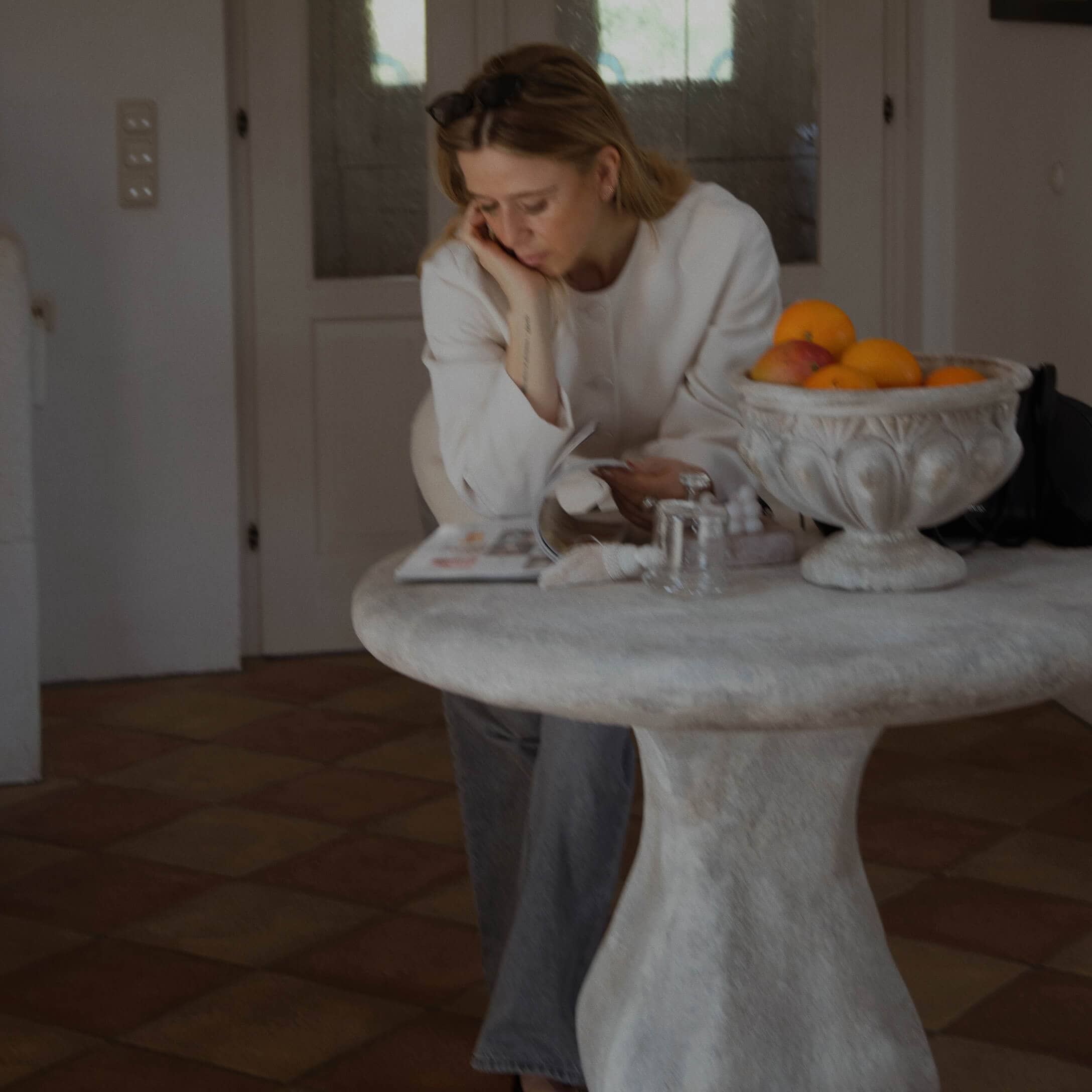 Frau im weissen Pullover liest an einem runden Steintisch mit Obstschale im Haus