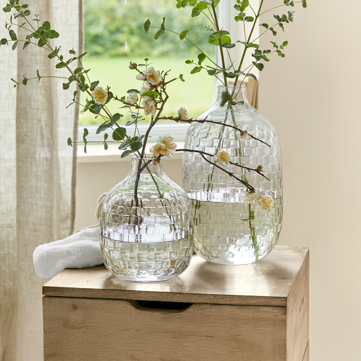 Vase 2er Set Qervilon Zwei durchsichtige Glasvasen mit Karomuster und grünen Zweigen mit Blüten auf dem Holztisch