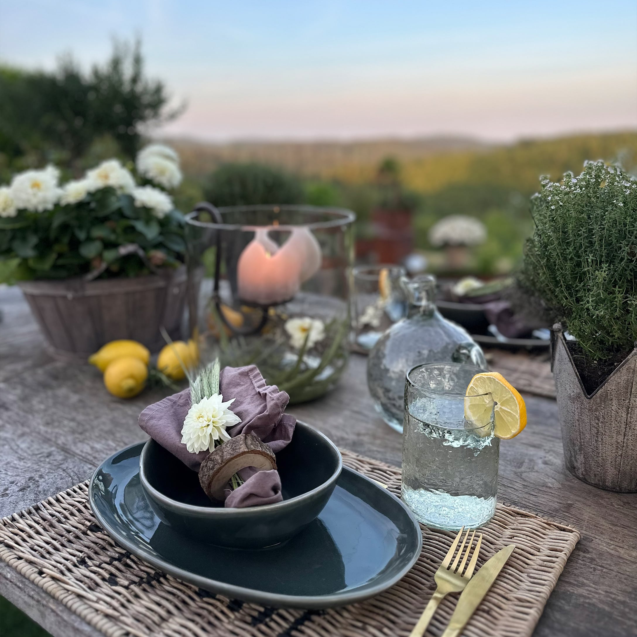 Rustikaler Tisch mit dunklem Keramikgeschirr, Leinenserviette und Glas Wasser mit Zitrone im Freien.