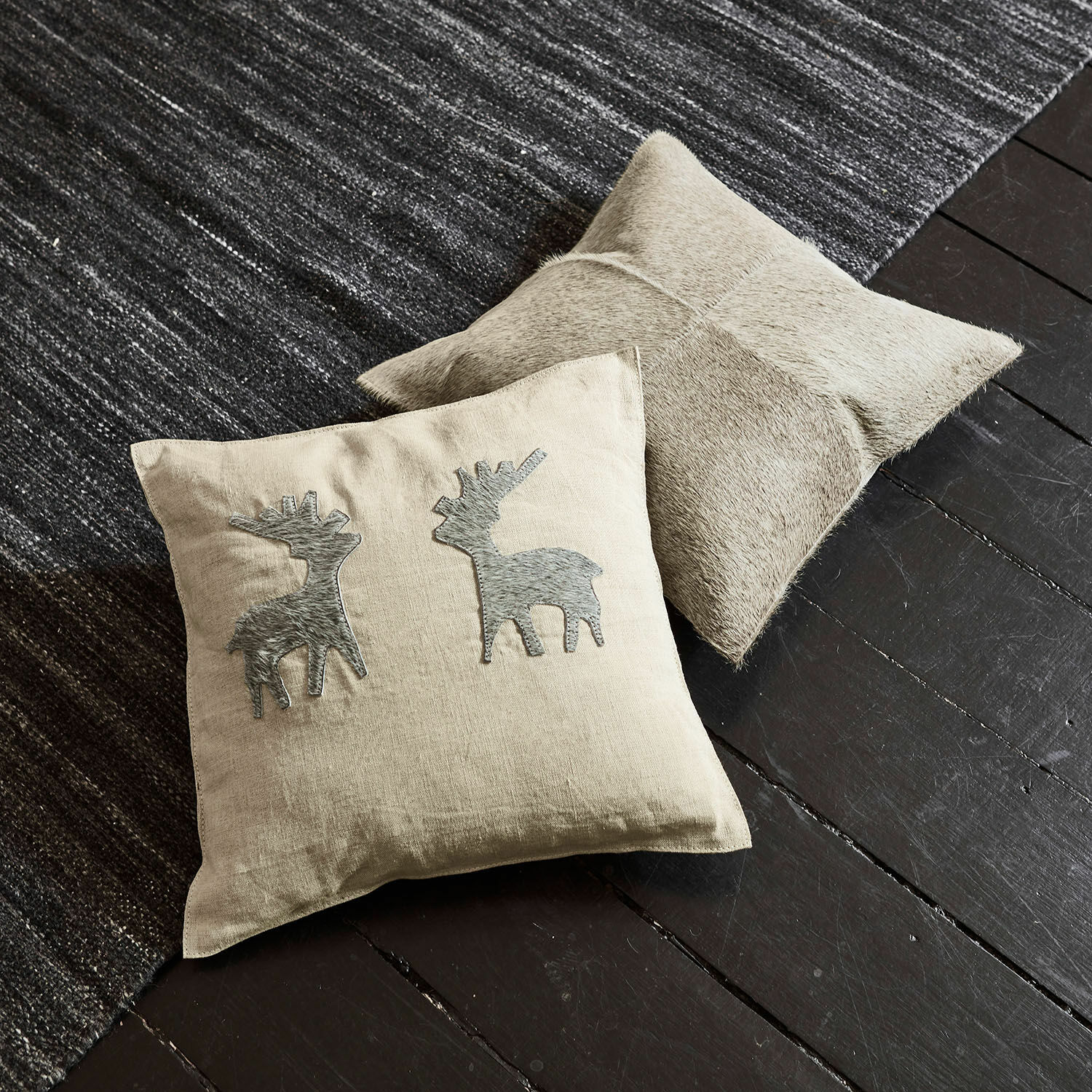 Kissenhülle 2er Set Beaupuy Beige Leinenkissen mit grauen Hirsch-Applikationen und hellbraunes Lederkissen auf dunklem Boden und Teppich.