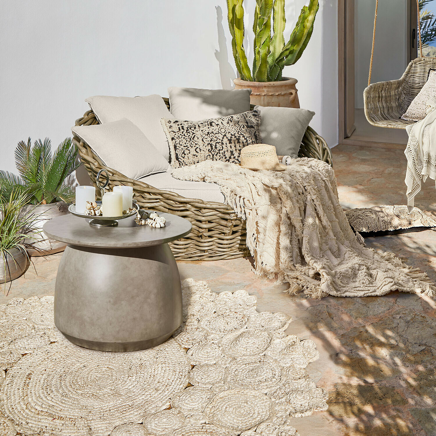 In- & Outdoor-Teppich Norlinn Ovales Rattansofa mit cremefarbenen Kissen und Decke auf Terrasse mit Beton-Couchtisch