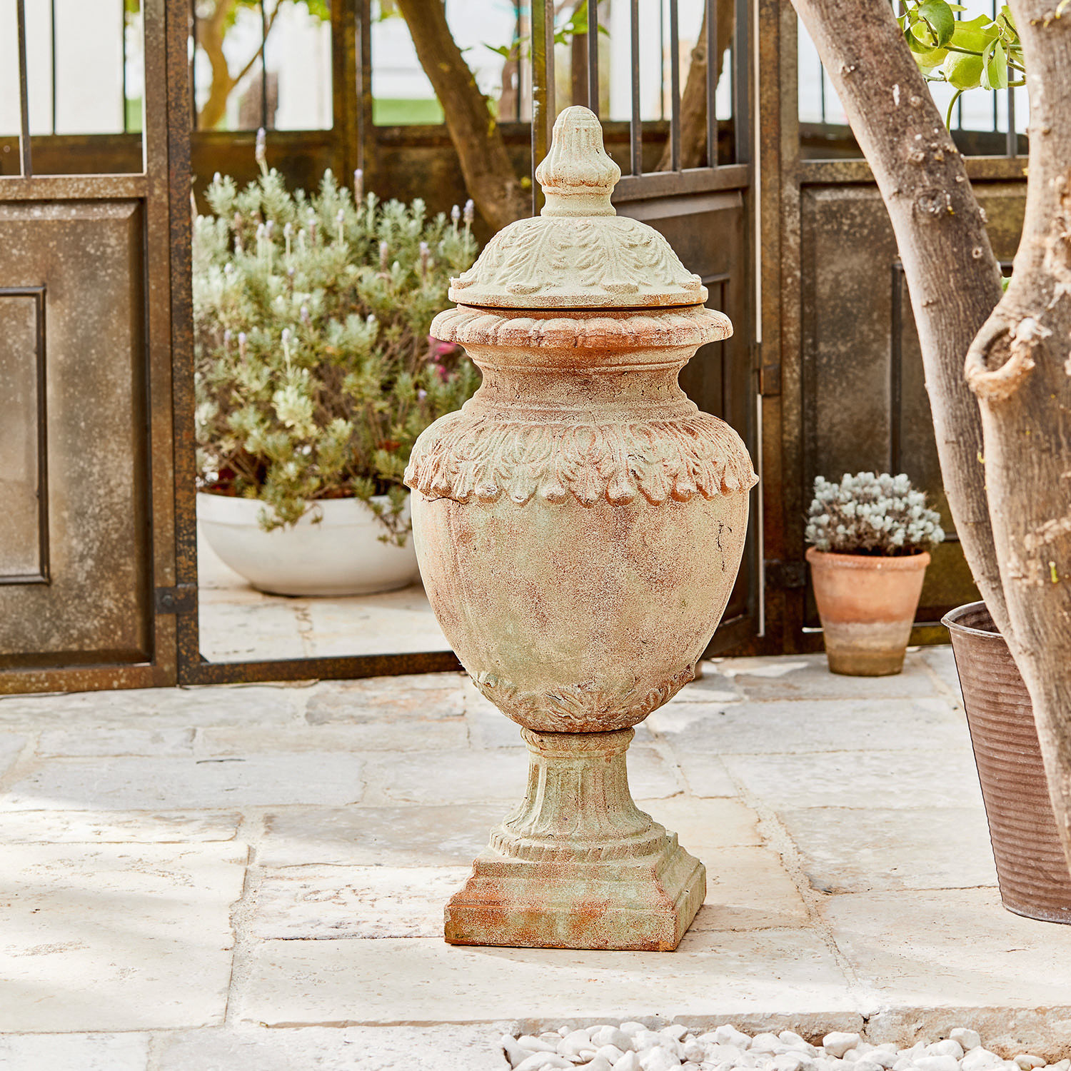 Deko-Objekt Persephone Dekorative Garten-Vase aus Terrakotta in Naturfarbe auf Steinboden mit Pflanzen im Hintergrund