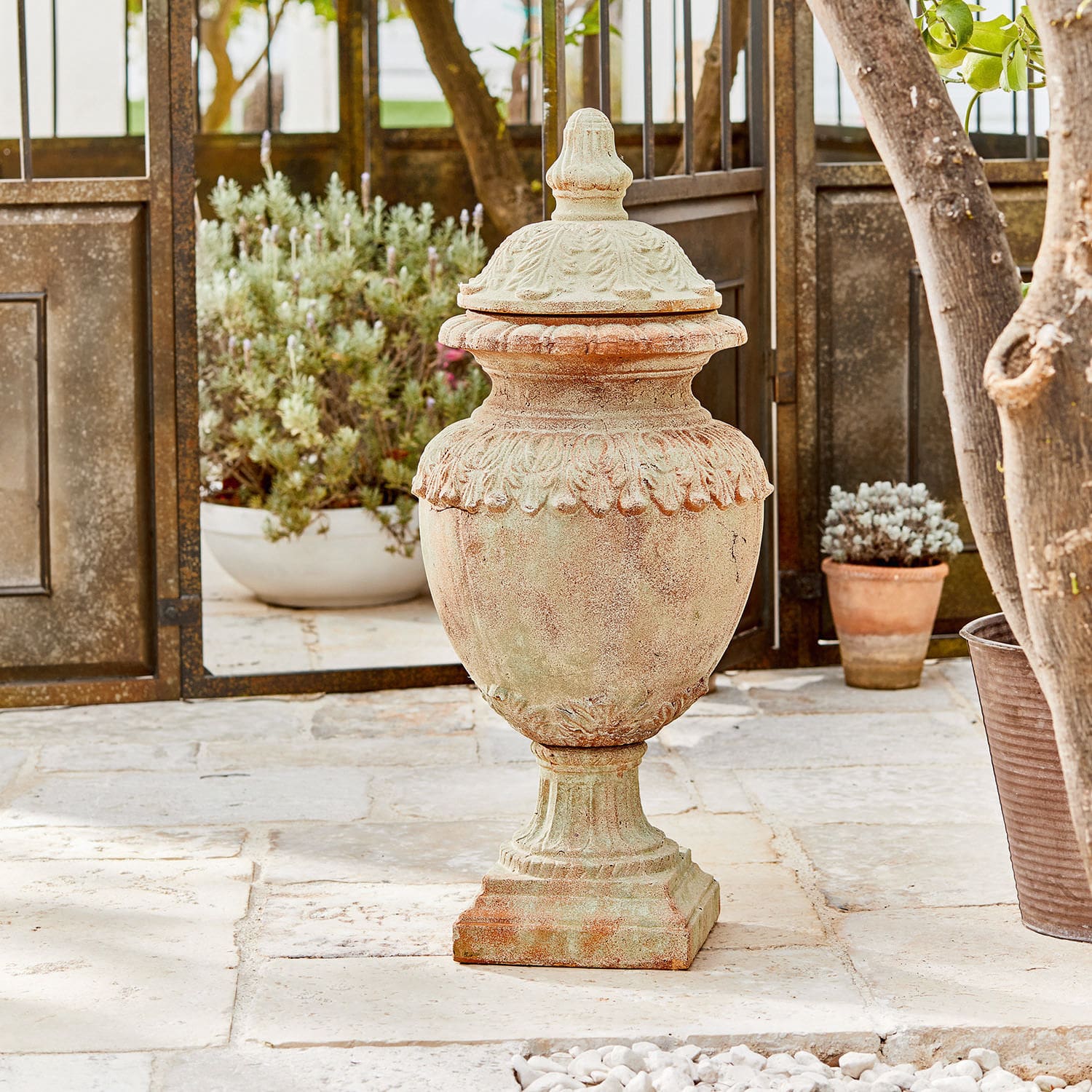 Deko-Objekt Persephone Dekorative Garten-Vase aus Terrakotta in Naturfarbe auf Steinboden mit Pflanzen im Hintergrund